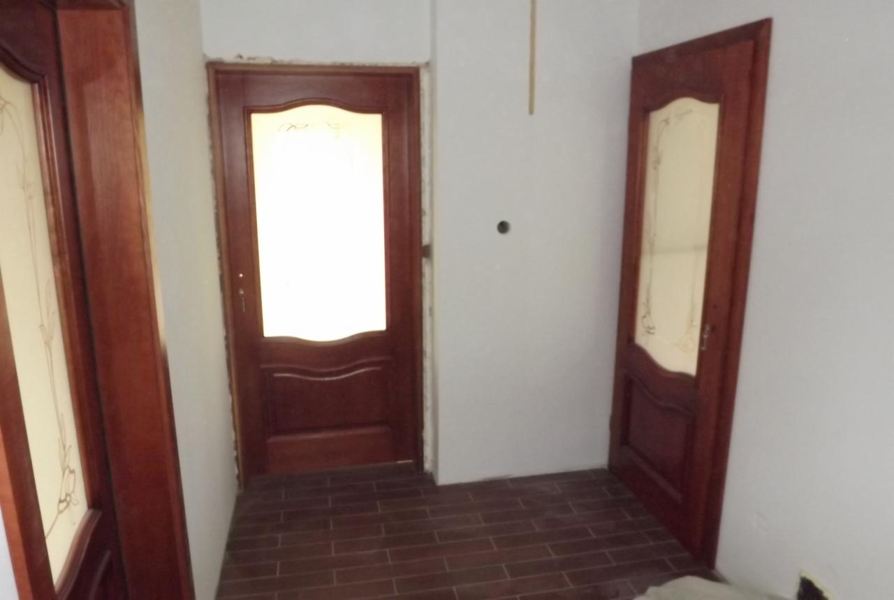 Wnętrze z trzema drewnianymi drzwiami z szybami, brązowa podłoga z płytek imitujących drewno, ściany w trakcie wykończenia.