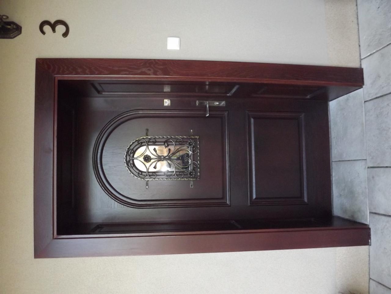 Ciemnobrązowe drzwi wejściowe z ozdobnym, metalowym wizjerem, osadzone w drewnianej ramie, z numerem domu '3' widocznym nad nimi.