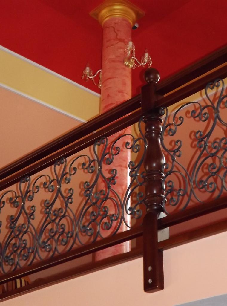 Fragment balustrady schodów z ciemnego drewna z ozdobnymi, kutymi elementami metalowymi na tle kolumny w stylu antycznym i czerwonego sufitu.