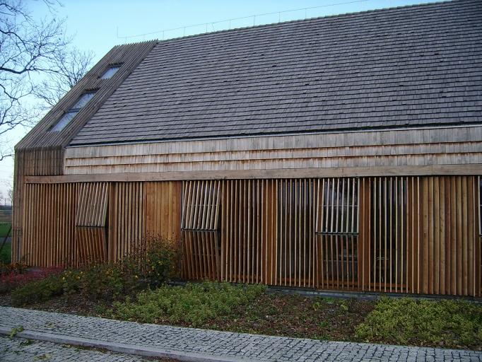 Nowoczesny budynek z drewnianą elewacją z pionowych listew i szarym dachem z gontów, widoczne otwarte okiennice z listew.