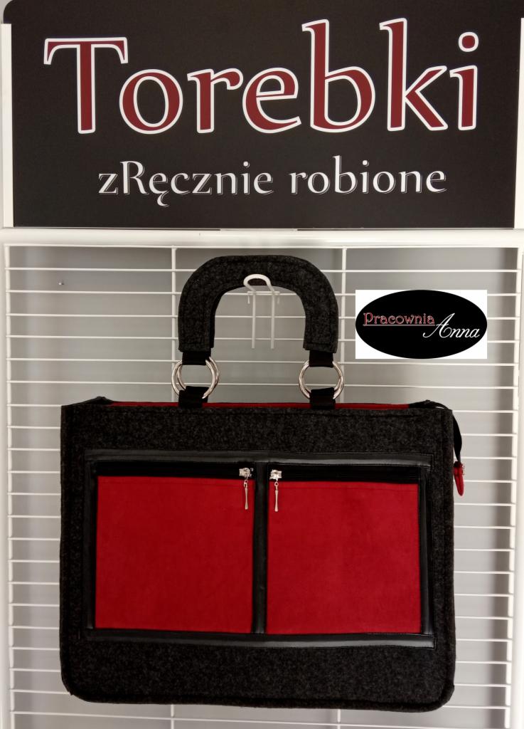 Szara filcowa torba z czerwonymi kieszeniami zapinanymi na zamek, wisząca na metalowym stojaku pod szyldem 'Torebki zręcznie robione' i logiem 'Pracownia Anna'.