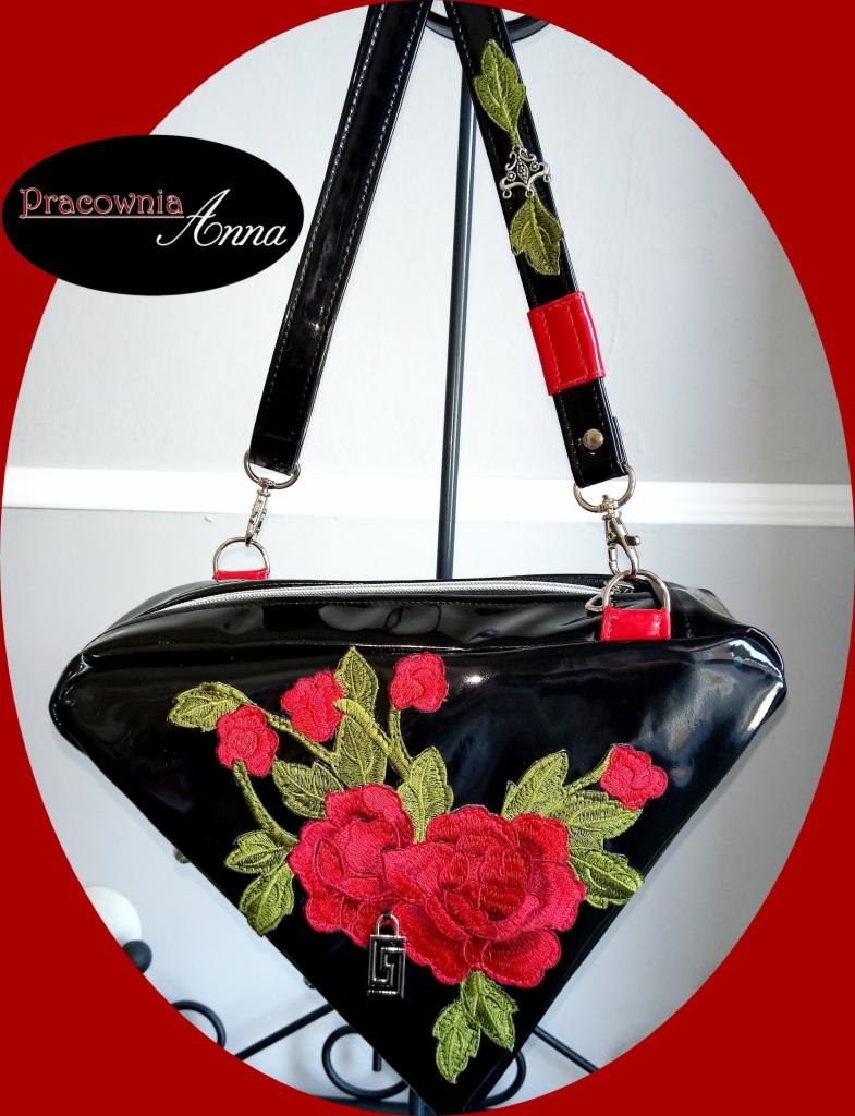 Czarna lakierowana torebka o trapezowym kształcie z haftowanymi czerwonymi różami i metalowym logo na tle jasnej ściany, zawieszona na stojaku.