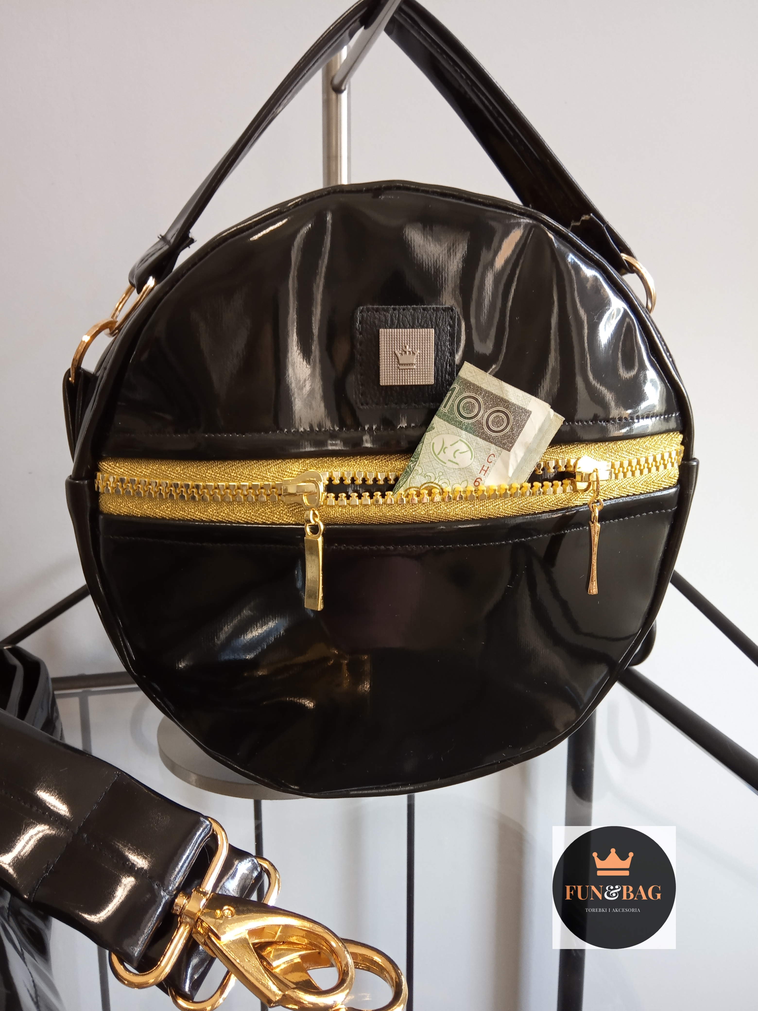 Lakierowana, okrągła, czarna torebka z złotym zamkiem, z wystającym banknotem 100 zł, prezentowana na stojaku.
