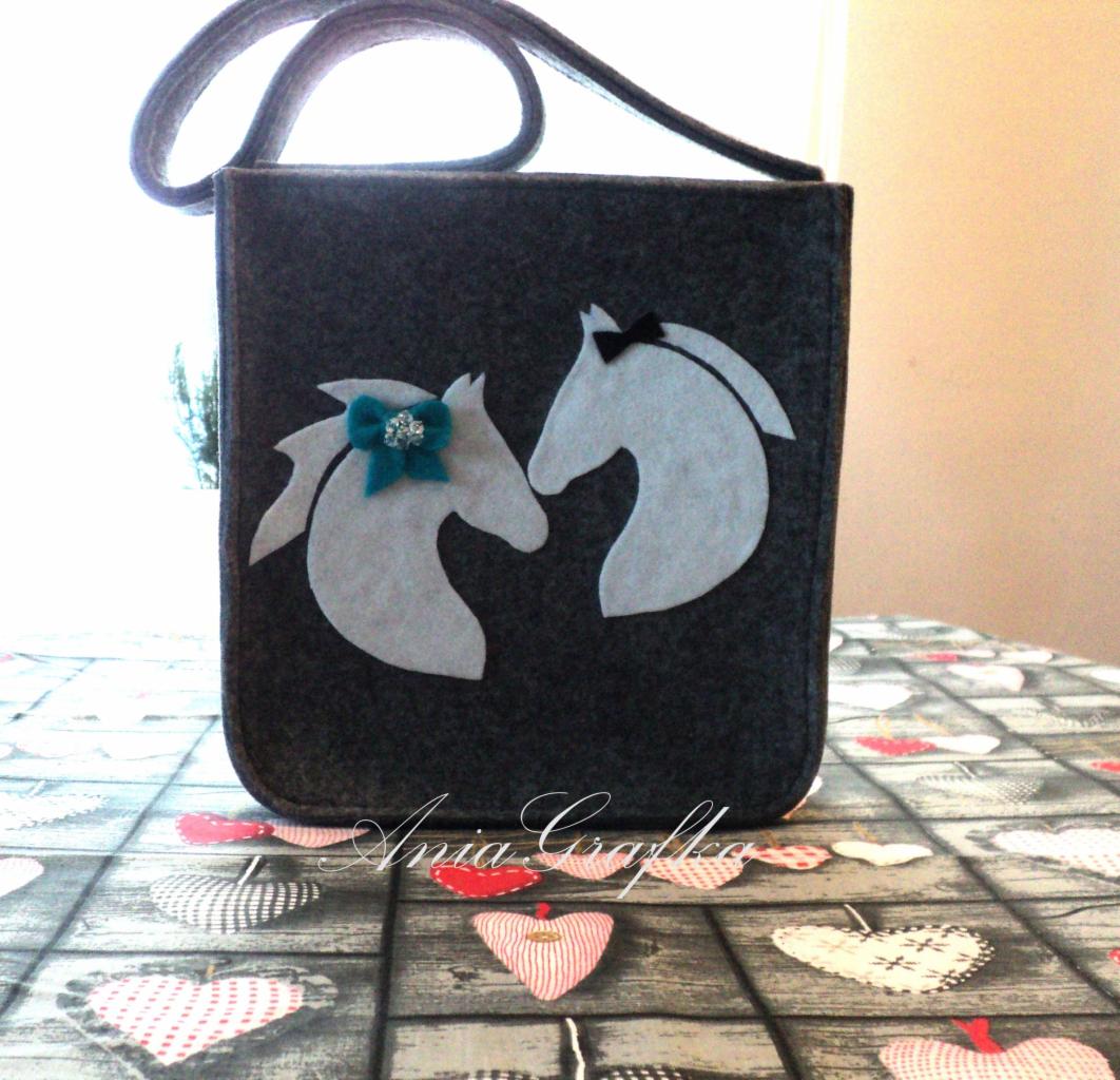 Szara filcowa torba z aplikacjami przedstawiającymi dwa białe końskie profile, jeden ozdobiony turkusową kokardką z kryształkami, drugi czarną kokardką, leżąca na stole z serduszkami.