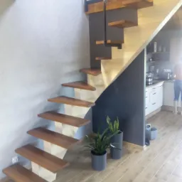 Nowoczesne schody wspornikowe z drewnianymi stopniami i metalową balustradą, prowadzące na antresolę, w tle widoczna kuchnia.