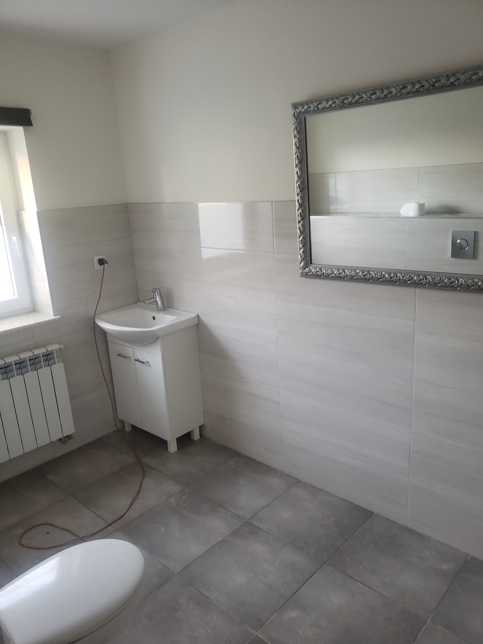 Wykończona łazienka z białą szafką pod umywalką, ozdobnym lustrem w srebrnej ramie, grzejnikiem i szarą podłogą z płytek.