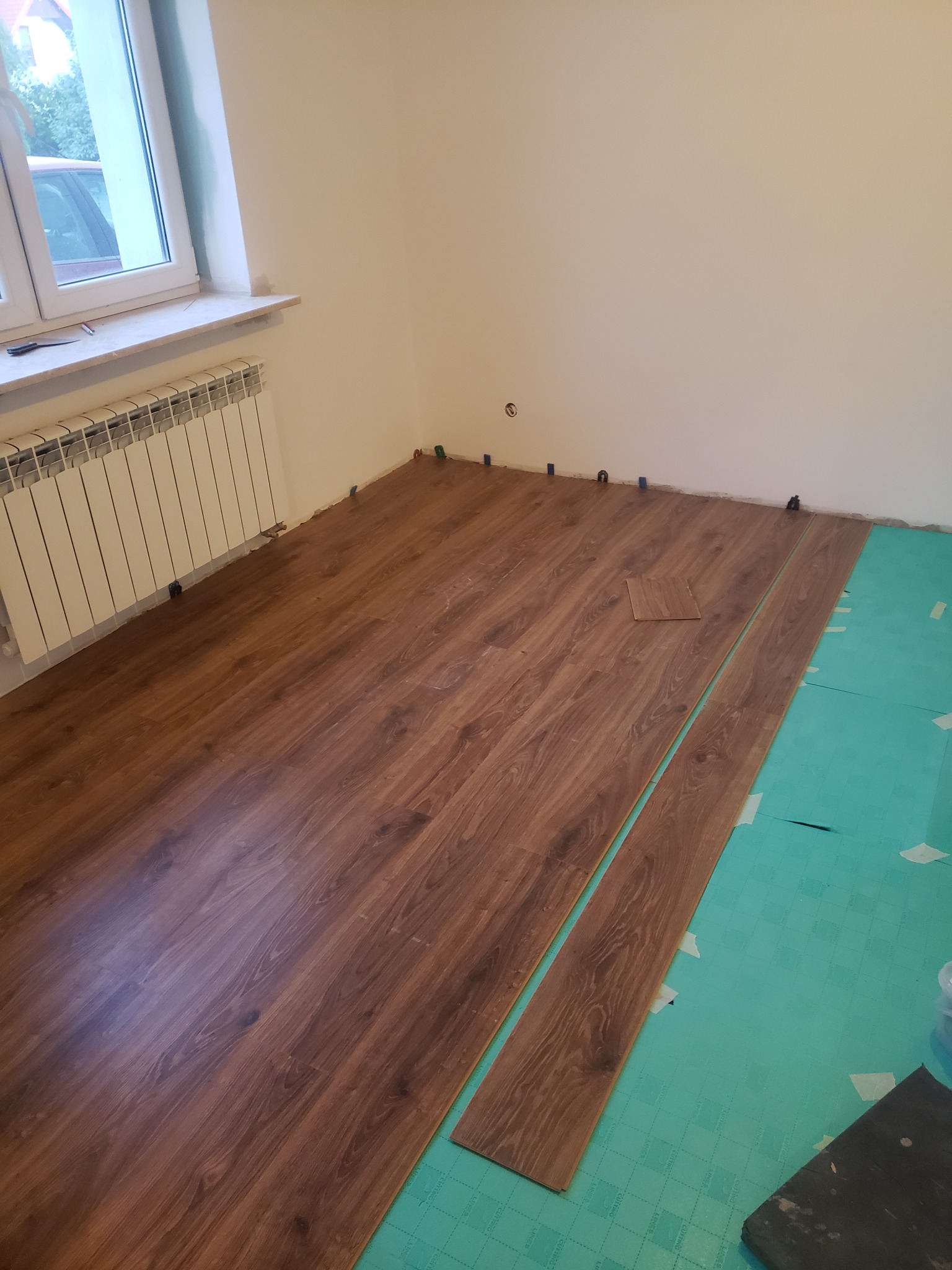 Układanie paneli podłogowych z brązowego drewna na zielonym podkładzie, widoczne kliny dystansowe przy ścianie i grzejnik.