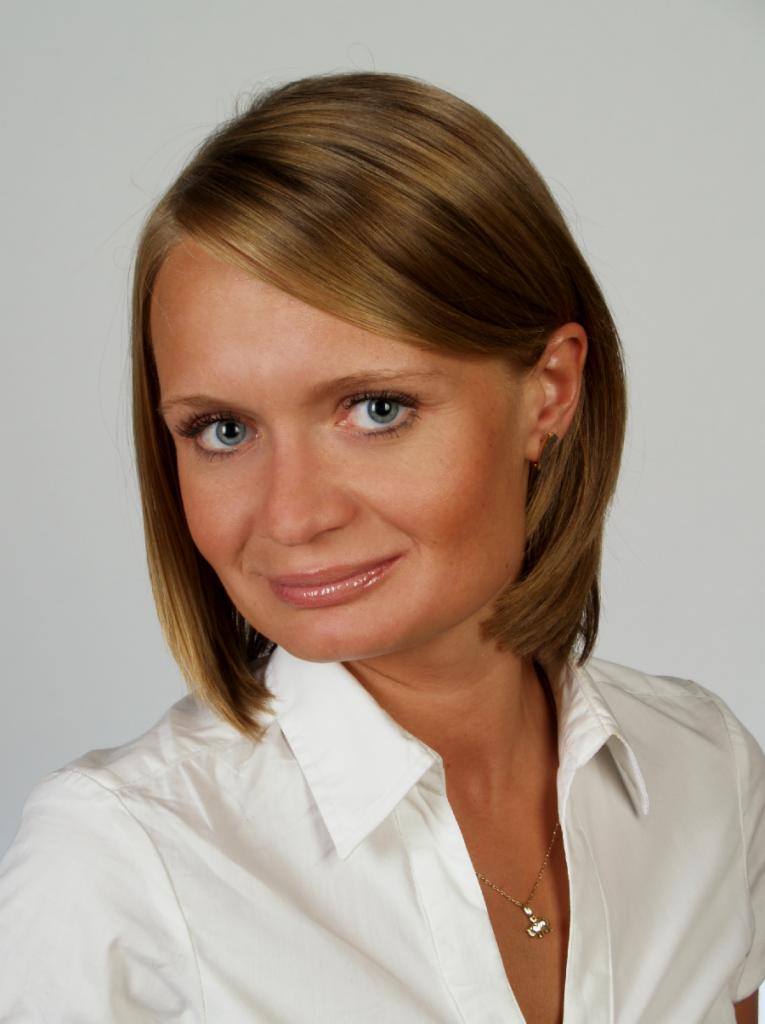 Portret kobiety w białej koszuli z delikatnym uśmiechem, złotym łańcuszkiem i kolczykami na jasnym tle.