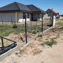 Nowoczesne ogrodzenie panelowe z podmurówką betonową, posesja z domem w trakcie budowy, widoczne prace ziemne.