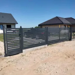 Nowoczesna, pozioma brama wjazdowa z metalu, pomalowana na szaro, z kulistymi zakończeniami słupków, na tle dwóch domów jednorodzinnych z ciemnymi dachami.