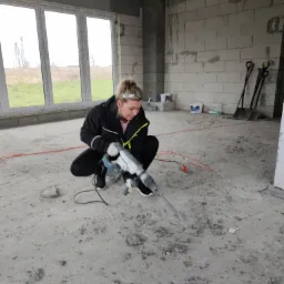 Kobieta w czarnej kurtce i białych rękawicach kuca i używa młota udarowego do kucia posadzki w surowym wnętrzu budynku w trakcie budowy. W tle okno, ściany z pustaków i narzędzia.