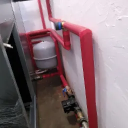 Szare naczynie przeponowe w systemie instalacji grzewczej z czerwonymi rurami izolowanymi termicznie, widoczne zawory i elementy przyłączeniowe, instalacja w pomieszczeniu z białą ścianą...