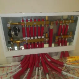 Rozdzielacz ogrzewania podłogowego Atech w białej obudowie, z czerwonymi rurami grzewczymi, widoczne białe rury na folii izolacyjnej z siatką.