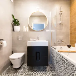 Nowoczesna łazienka z szarą ceramiką, czarną szafką pod umywalką, wzorzystą podłogą, okrągłym lustrem z drewnianą ramą i wanną z parawanem oraz dekoracyjnymi świecami i kieliszkiem wina.