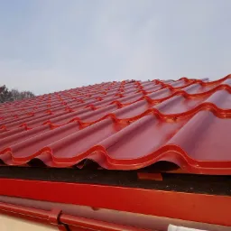 Czerwona blachodachówka modułowa ułożona na dachu, widoczna rynna w tym samym kolorze, w tle fragment drzewa i jasne niebo.