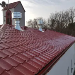 Czerwona blachodachówka z dwoma metalowymi kominami wentylacyjnymi na tle wieży z megafonami i nagich drzew