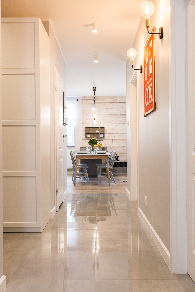 Wnętrze mieszkania z perspektywy korytarza: błyszcząca, jasna podłoga, białe ściany z dekoracyjnym napisem 'Everything OK', w tle stół z krzesłami i drewniana ściana z półkami.