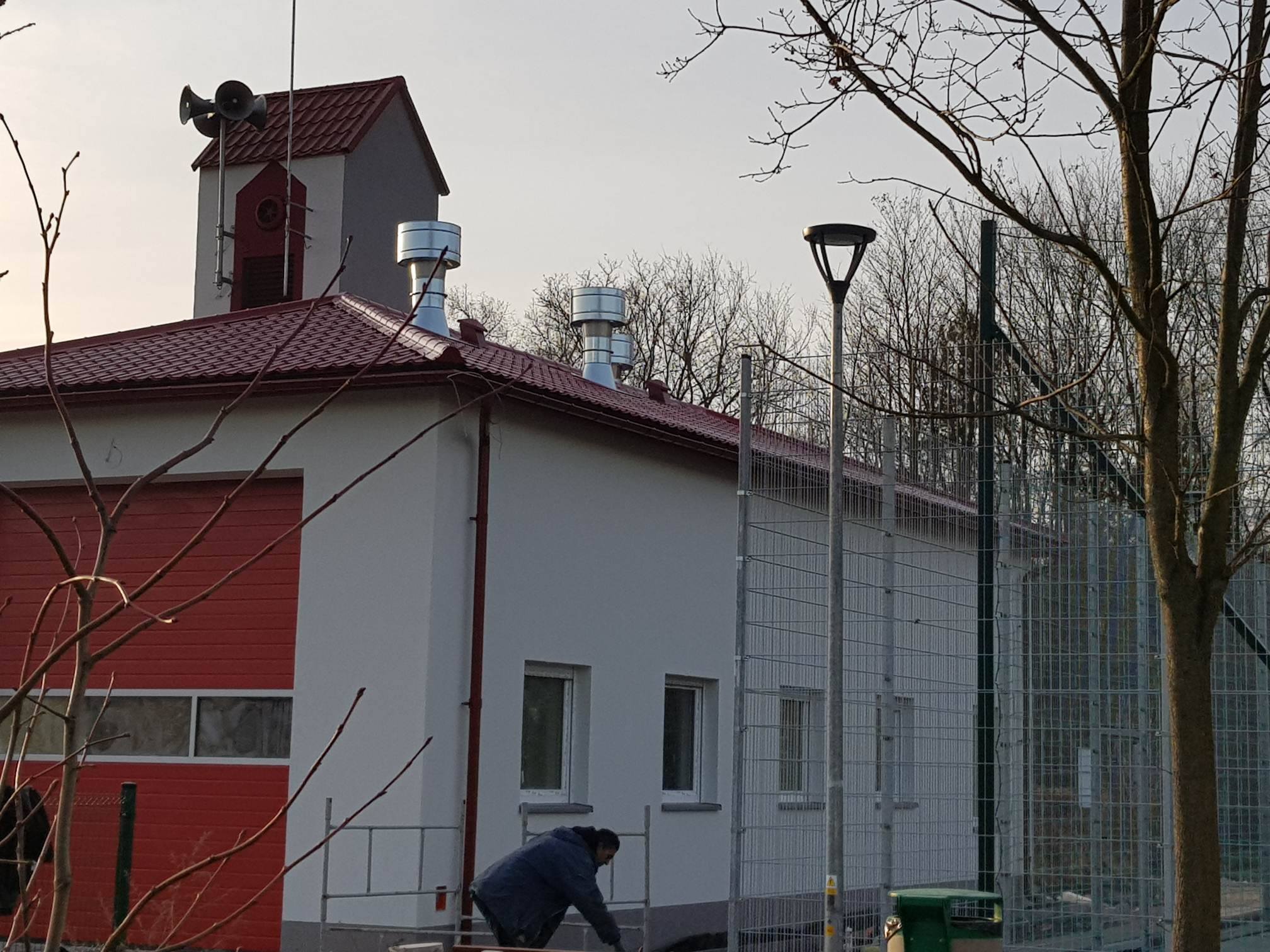 Pracownik w niebieskiej kurtce maluje elewację budynku z czerwoną bramą garażową i dachem z dachówki, widoczne metalowe kominy i wieżyczka z głośnikami na dachu.