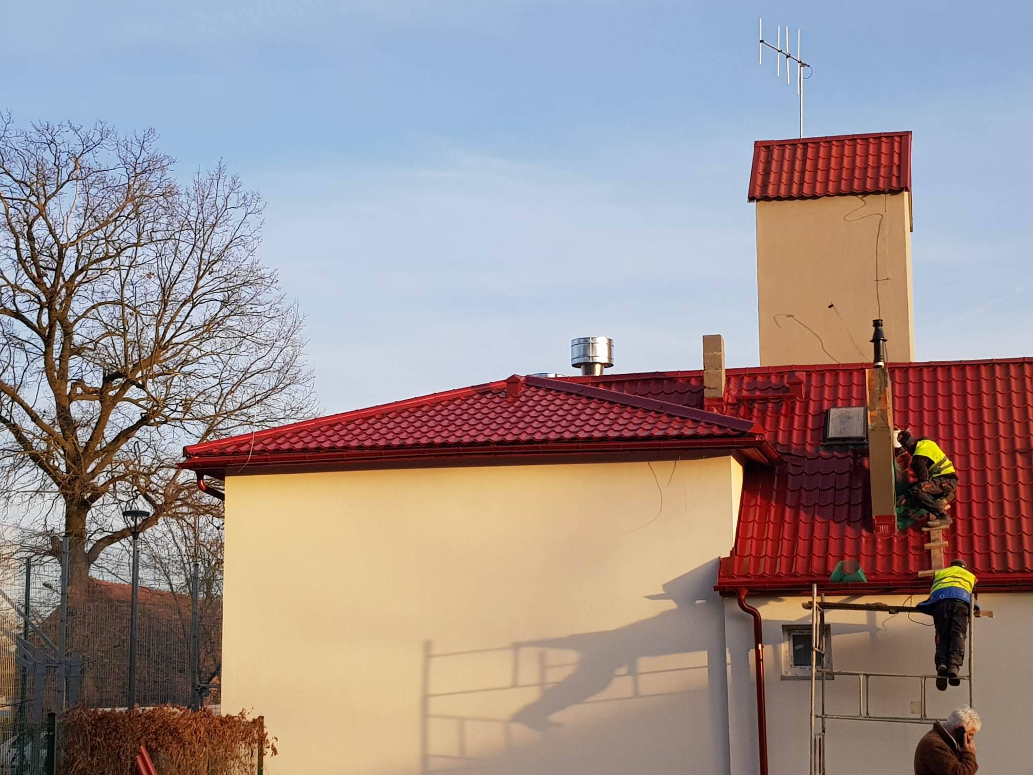 Dwóch pracowników na rusztowaniu montuje element na dachu budynku z czerwoną dachówką, widoczny komin z anteną i drzewo bez liści w tle.