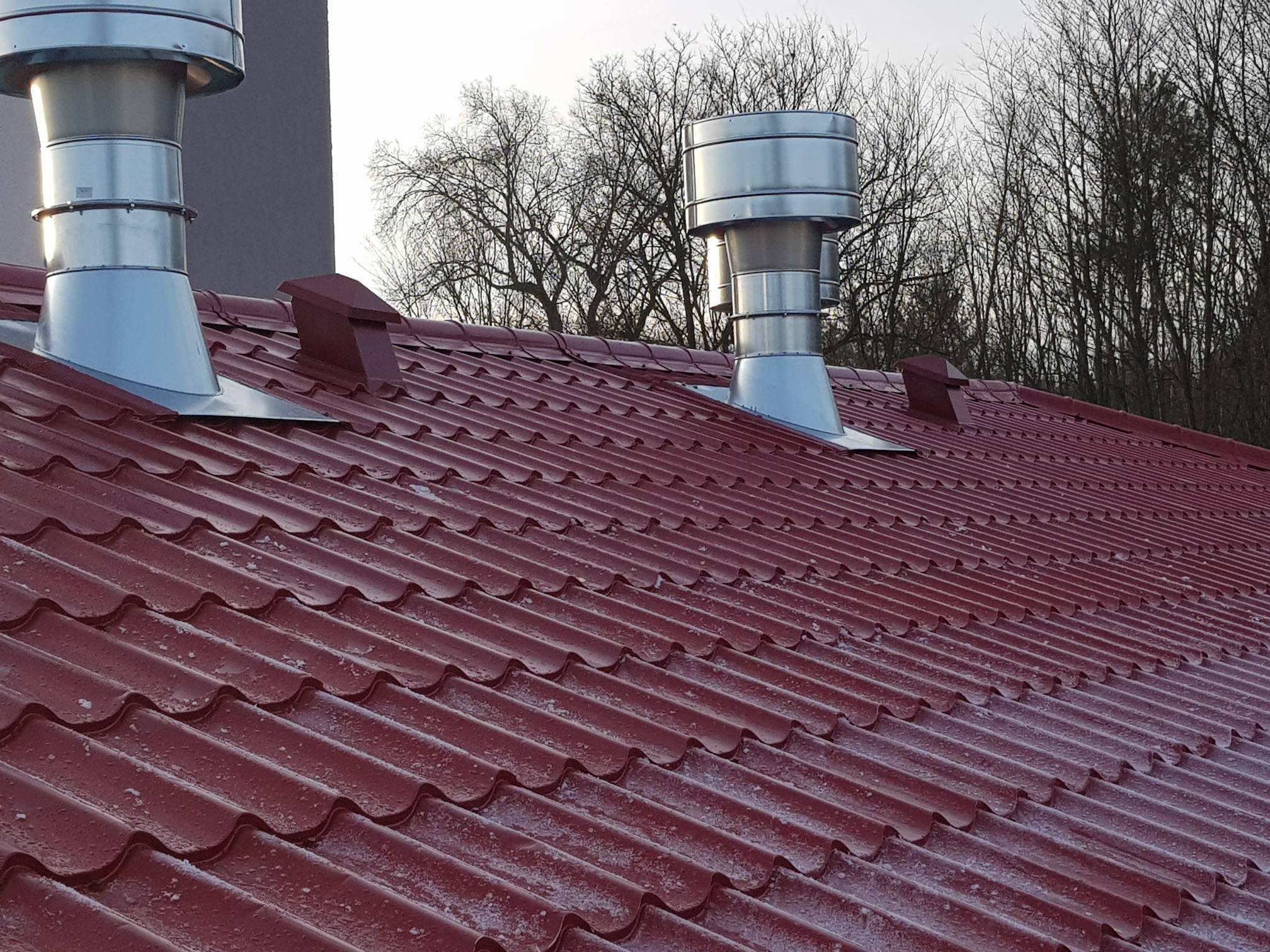 Czerwony dach z blachy trapezowej z dwoma metalowymi kominami wentylacyjnymi i małymi wywietrznikami dachowymi, widoczne drzewa bez liści w tle.