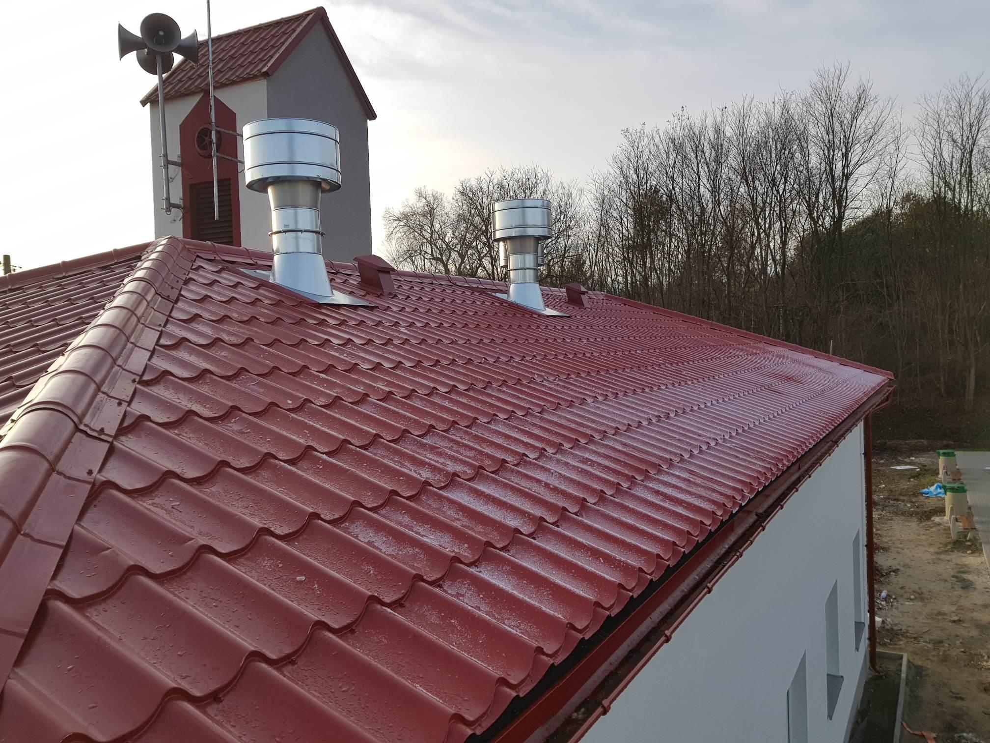 Czerwona blachodachówka z dwoma metalowymi kominami wentylacyjnymi na tle wieży z megafonami i nagich drzew