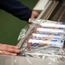 Ręka z obrączką przy pakowaniu w folię termokurczliwą zestawu puszek na linii produkcyjnej.