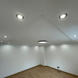 Pomalowane na biało wnętrze z drewnianą podłogą i oświetleniem sufitowym, w tym reflektory punktowe i centralna lampa, z widoczną instalacją rurową przy suficie.