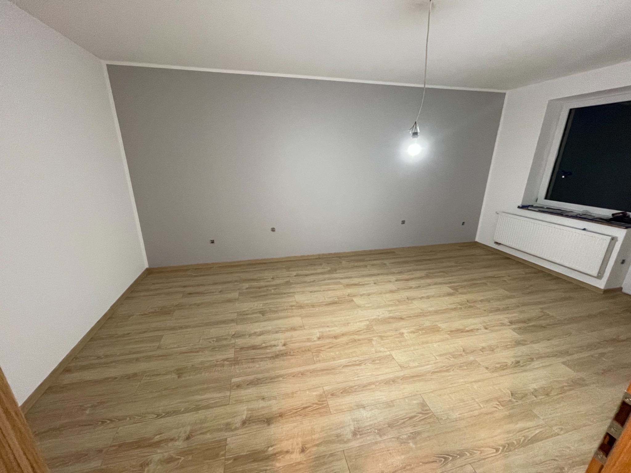 Pomalowane na szaro ściany w pustym pokoju z drewnianą podłogą, widoczny grzejnik pod oknem i wisząca żarówka na kablu.