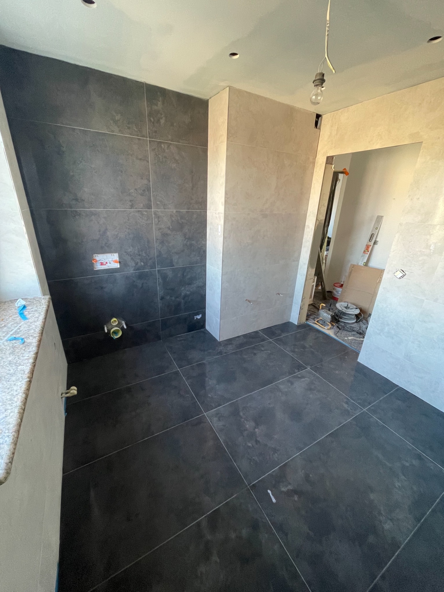 Ciemne płytki podłogowe i częściowo wyłożone ściany w trakcie układania w łazience, widoczne przyłącza wodne i narzędzia w tle.