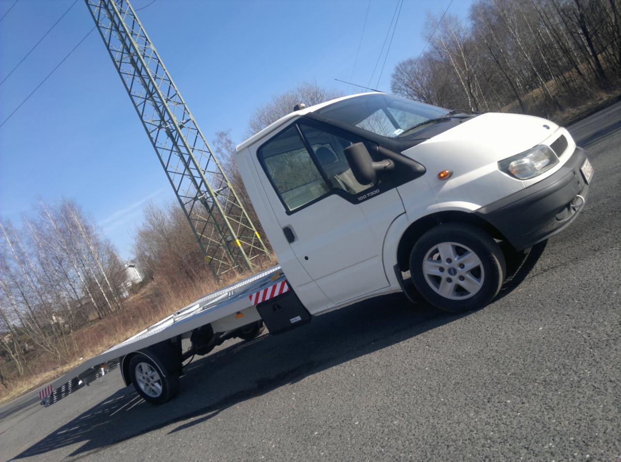Biała laweta Ford Transit 100 T300 na asfalcie, z widoczną konstrukcją stalową wysokiego napięcia w tle.