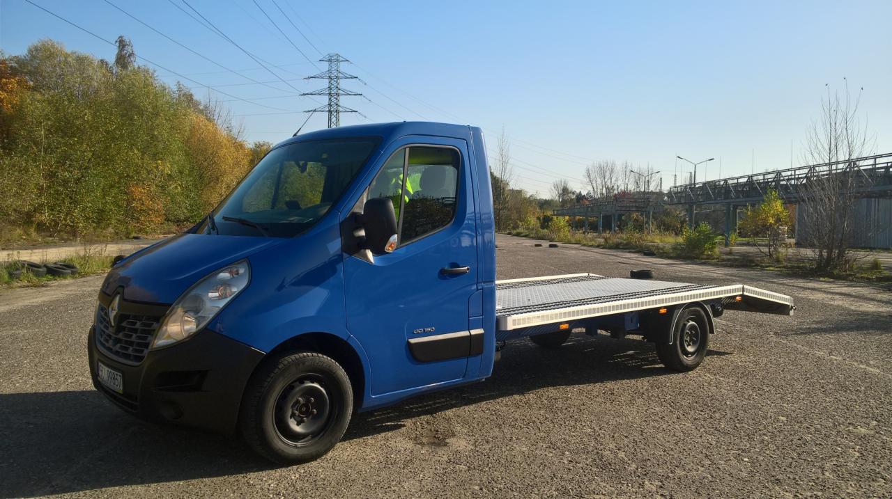Niebieska laweta Renault Master z platformą transportową na szarym placu, w tle widoczne drzewa i konstrukcja mostu.