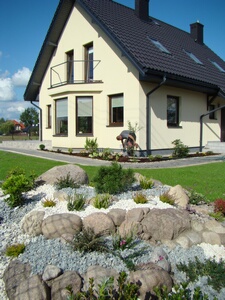 Ułożony z kamieni i żwiru skalniak z roślinami na pierwszym planie, w tle dom jednorodzinny z zadbanym trawnikiem i pracującą osobą, słoneczny dzień.
