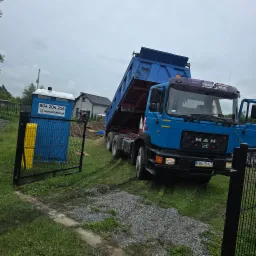 Niebieska ciężarówka MAN wyładowuje piasek na posesji obok przenośnej toalety TOI TOI. Widoczne świeże ślady opon na trawie.
