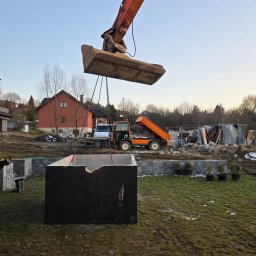Damian Nagórski - Montaż betonowego zbiornika na posesji z użyciem koparki. W tle pomarańczowa wywrotka i częściowo widoczny dom. Etap prac ziemnych.