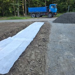 Damian Nagórski - Wykop z białą geowłókniną, obok hałda żwiru i niebieska wywrotka na tle lasu. Prace ziemne w toku, przygotowanie podłoża.