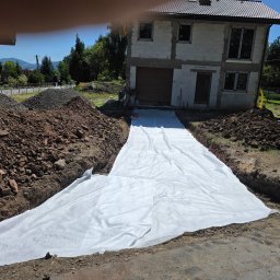 Damian Nagórski - Przygotowanie podłoża pod nawierzchnię: wykop z geowłókniną na tle domu w budowie, hałdy ziemi i żwiru. Słoneczny dzień.