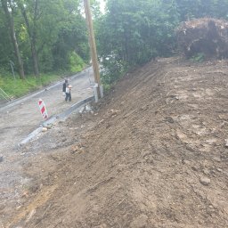 Damian Nagórski - Budowa drogi gruntowej z krawężnikami, widoczny fragment korzenia drzewa. Dwie osoby spacerują po nowej drodze. Prace ziemne w toku, teren nierówny.