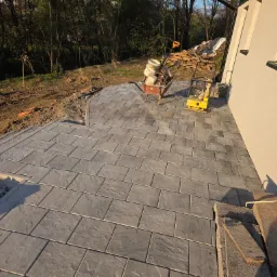 Ułożona szara kostka brukowa na tarasie domu, z widocznymi narzędziami (taczka, ubijarka) i materiałami budowlanymi w tle, w słoneczny dzień.