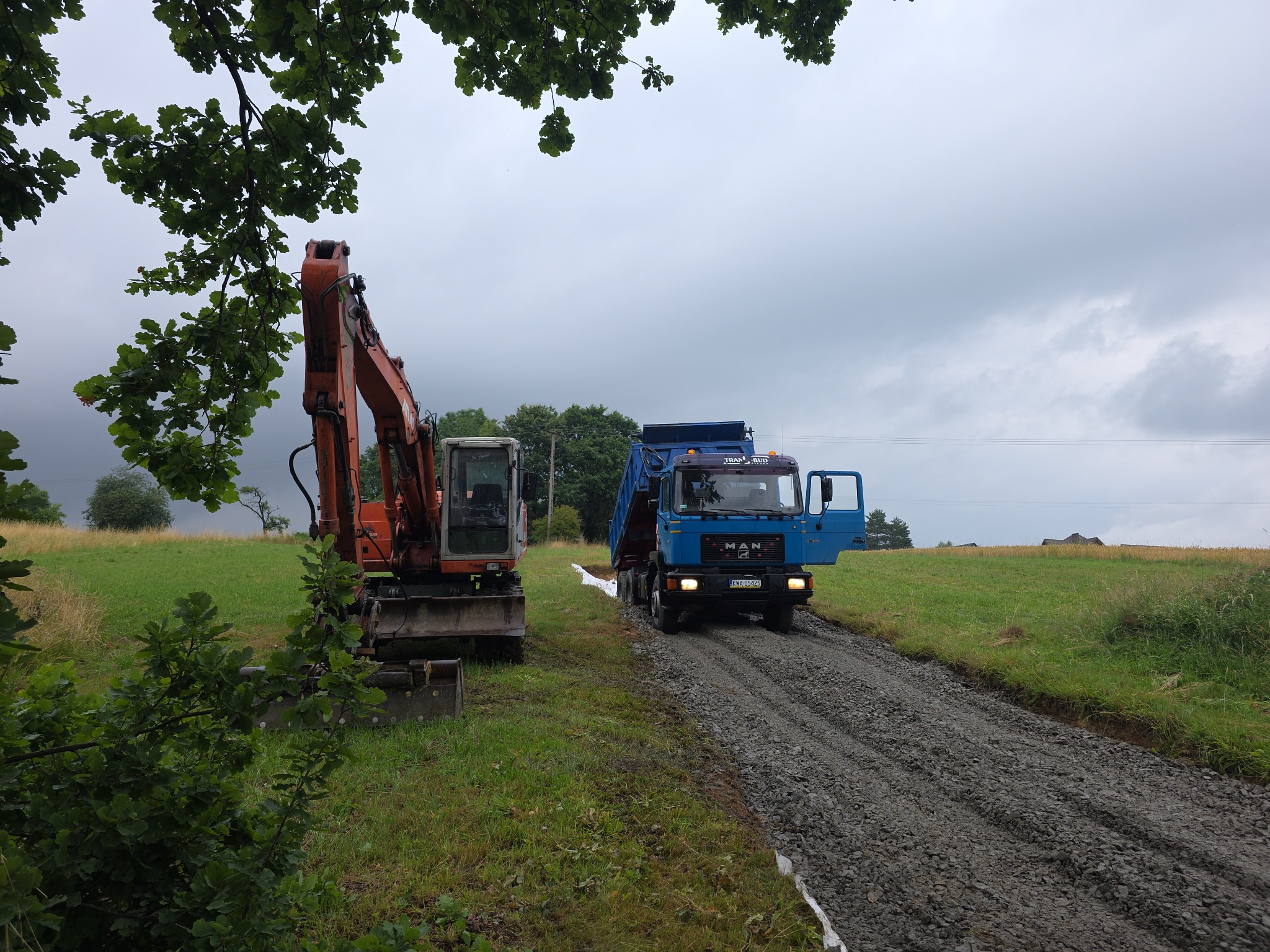 Niebieska ciężarówka MAN wysypuje żwir na utwardzoną drogę polną obok pomarańczowej koparki na tle zielonego pola i pochmurnego nieba.