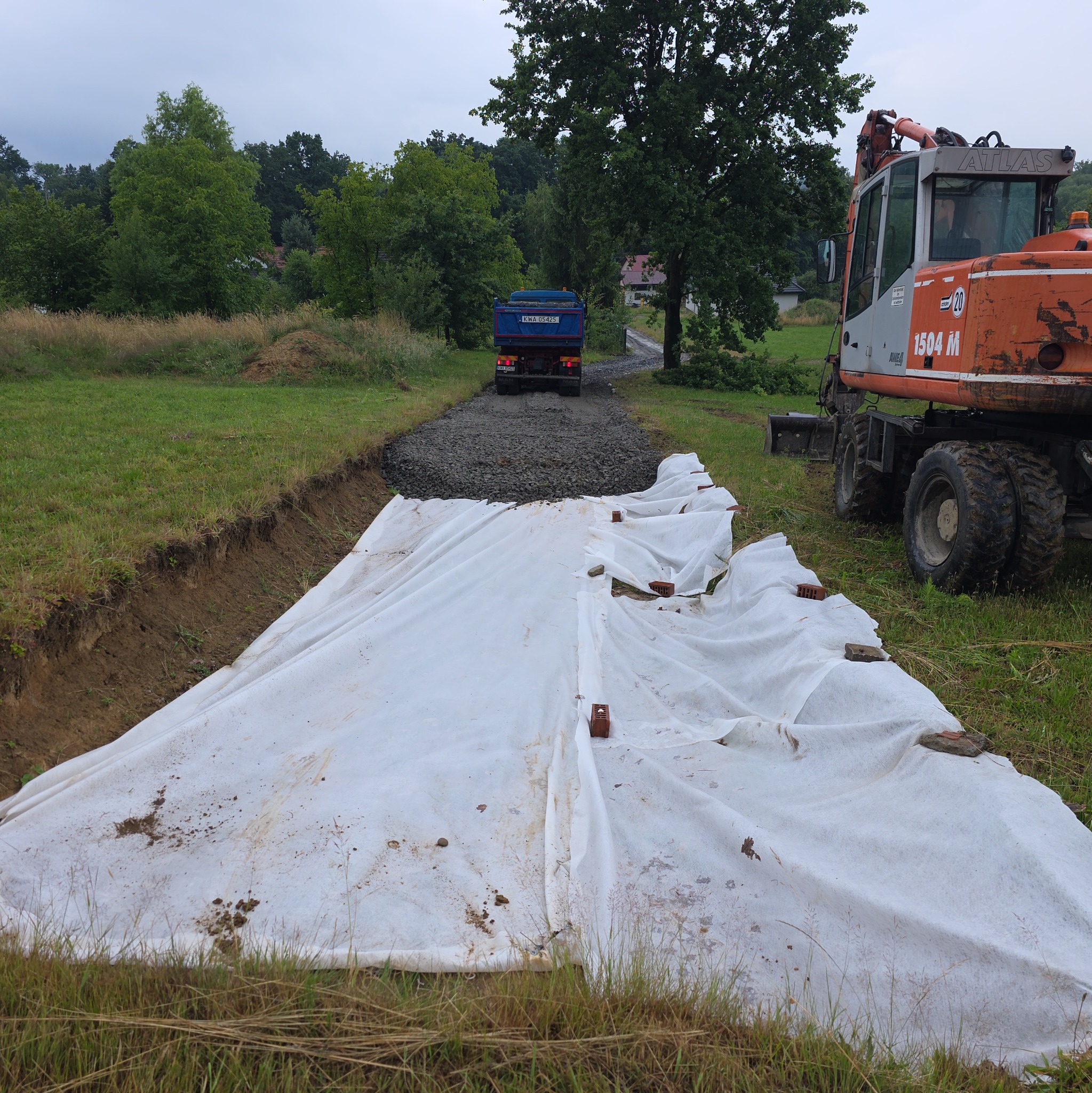 Przygotowanie podłoża pod drogę w Cieszynie: wykop, geowłóknina zabezpieczona cegłami, żwir i ciężarówka w tle. Koparka obok.