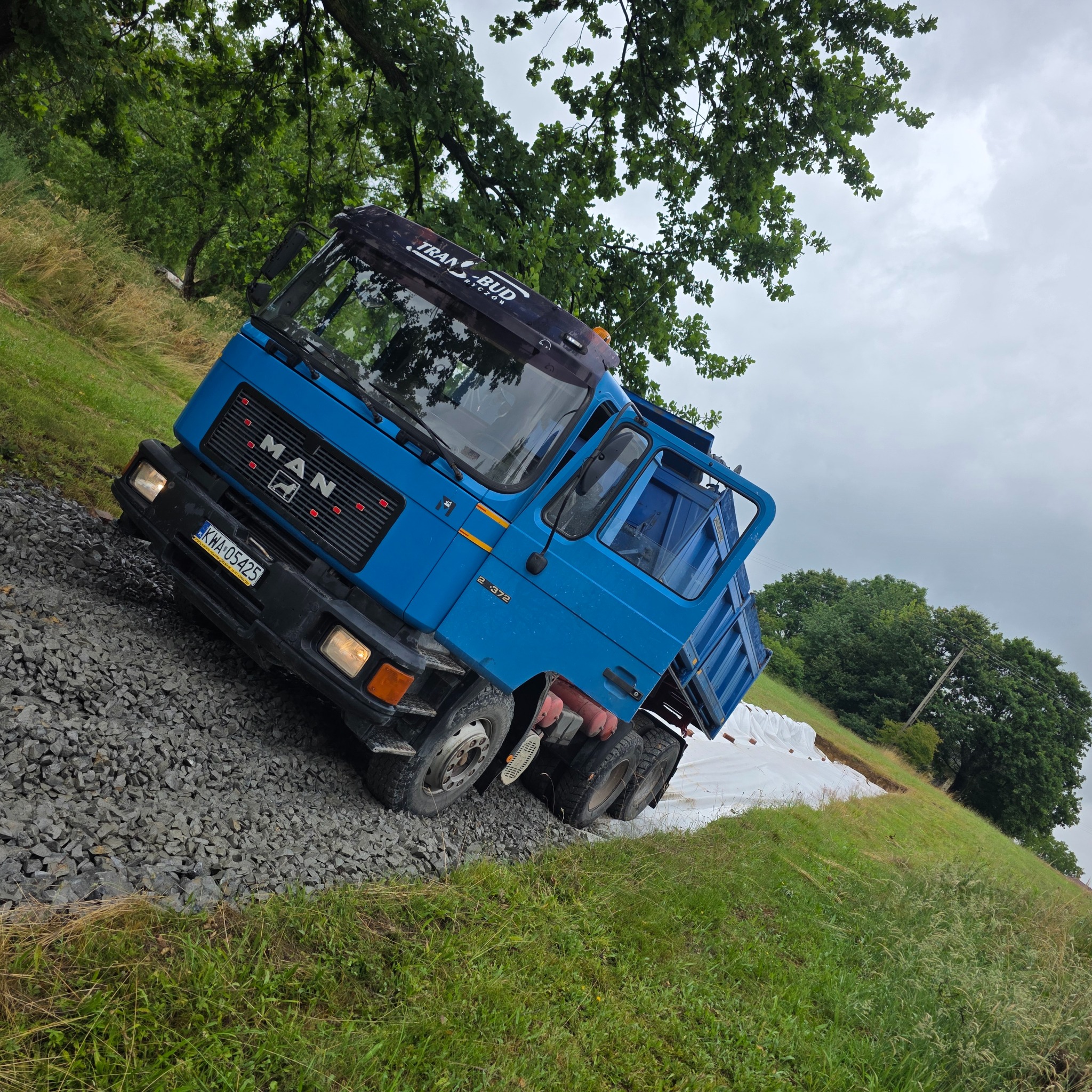 Niebieski samochód ciężarowy MAN na żwirowej drodze w pochmurny dzień, z widocznymi workami z materiałem budowlanym w tle.