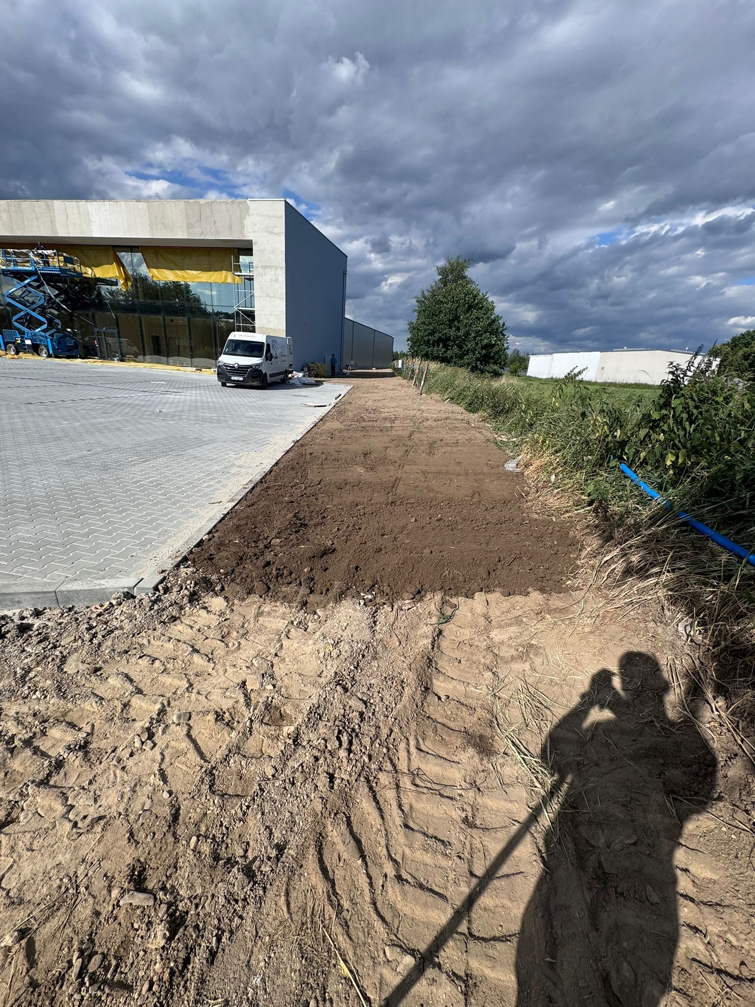 Przygotowany teren pod ścieżkę obok parkingu z kostki, w tle budynek i van. Widoczny cień osoby robiącej zdjęcie, podkreślający etap prac ziemnych.