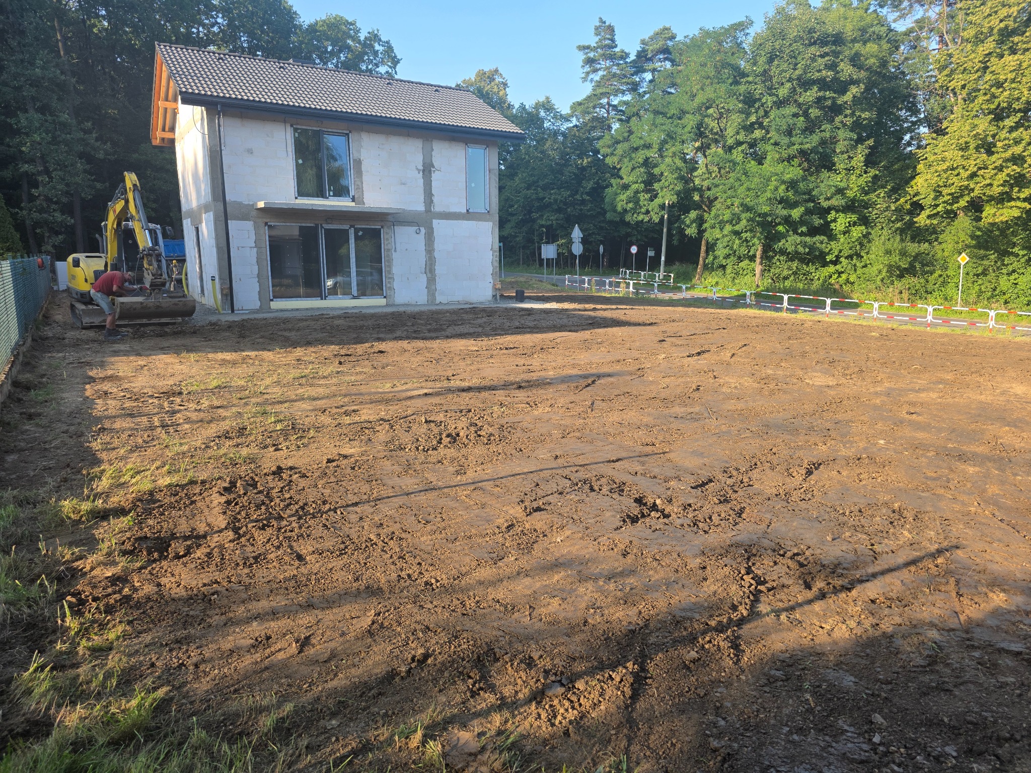 Żółta koparka przy białym domu z dwuspadowym dachem, wyrównany teren, w tle zieleń drzew. Przygotowanie terenu pod przyszły ogród.