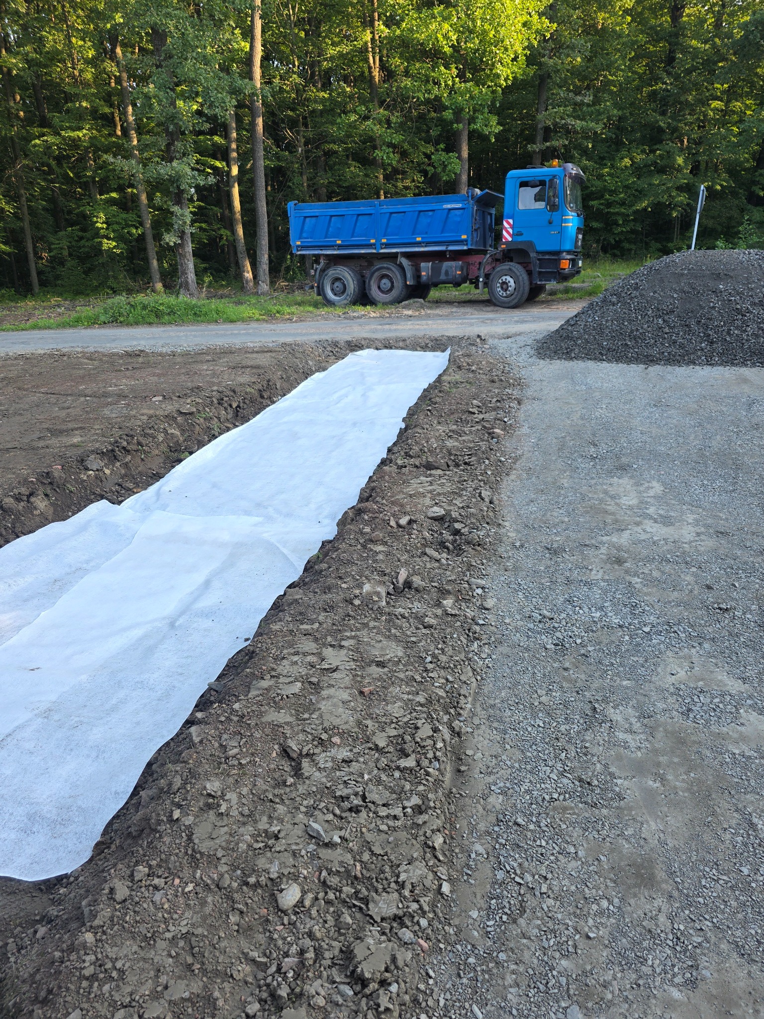 Wykop z białą geowłókniną, obok hałda żwiru i niebieska wywrotka na tle lasu. Prace ziemne w toku, przygotowanie podłoża.