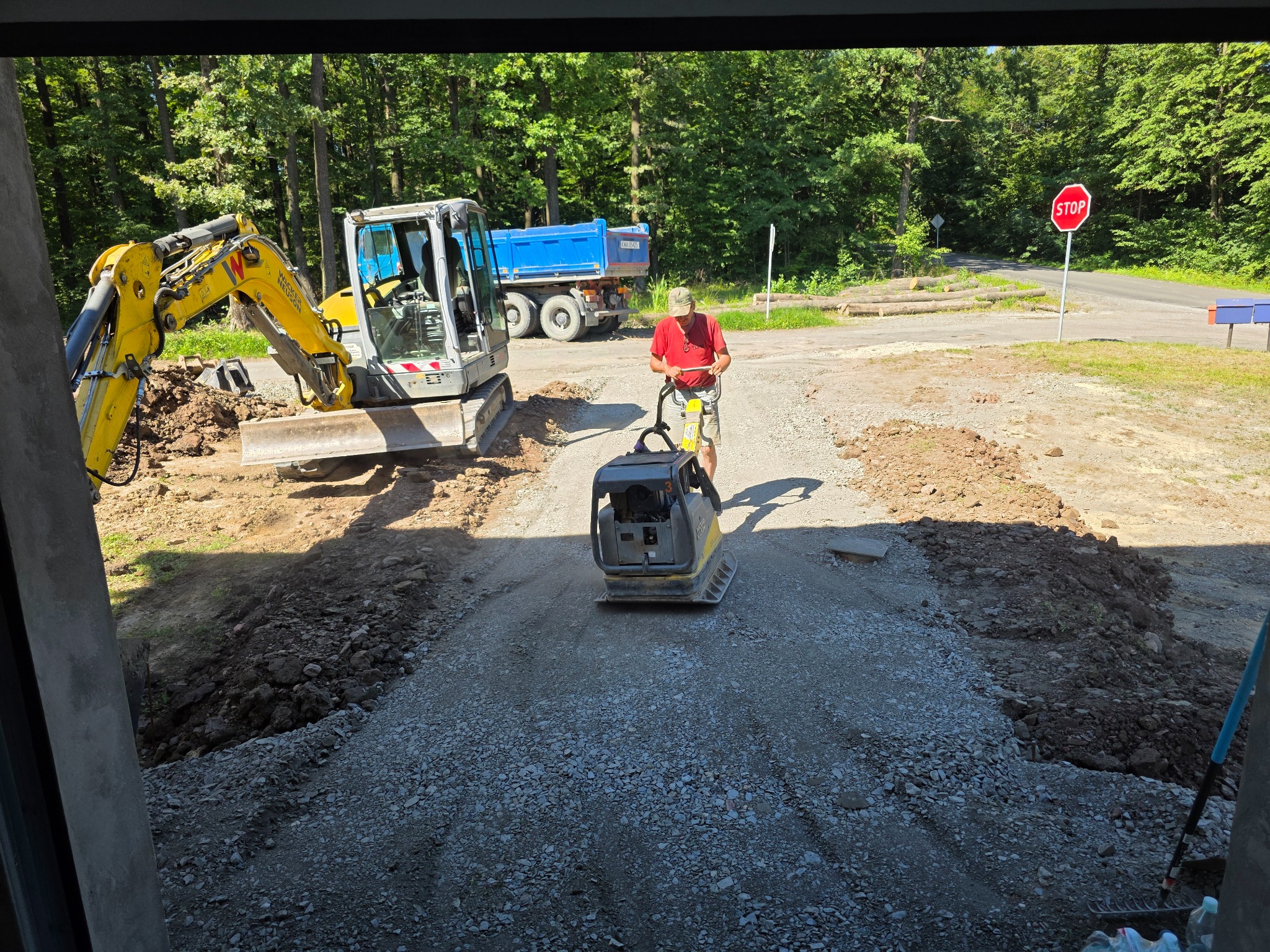 Prace ziemne w toku: ubijarka wibruje żwirowe podłoże drogi, w tle koparka i wywrotka. Widok z wnętrza budynku na teren robót.