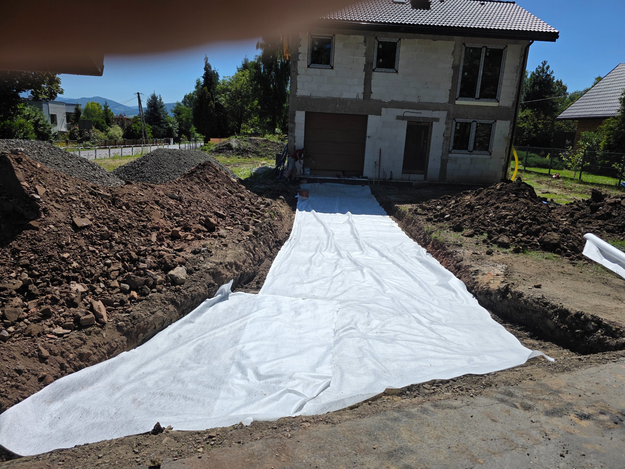 Przygotowanie podłoża pod nawierzchnię: wykop z geowłókniną na tle domu w budowie, hałdy ziemi i żwiru. Słoneczny dzień.