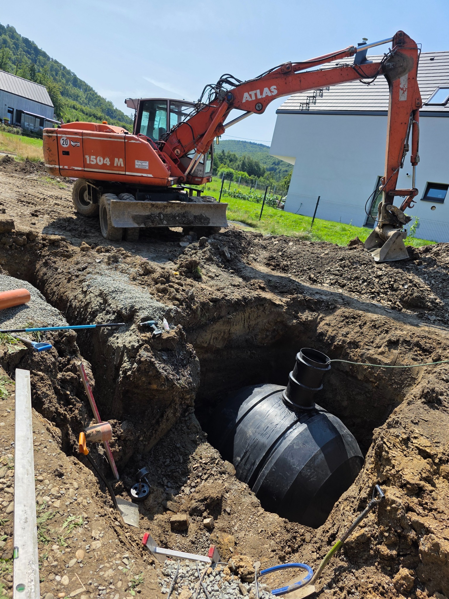 Montaż zbiornika retencyjnego w wykopie z użyciem koparki Atlas 1504 M. Widoczne narzędzia i elementy instalacji. Krajobraz z budynkami w tle.