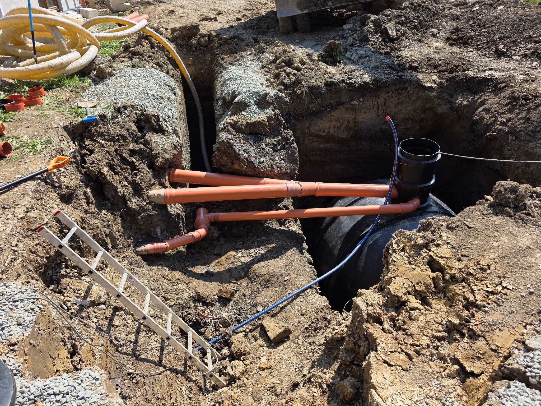 Czarny zbiornik w wykopie z rurami drenażowymi i warstwą żwiru, widoczne narzędzia i drabina, teren po niwelacji.