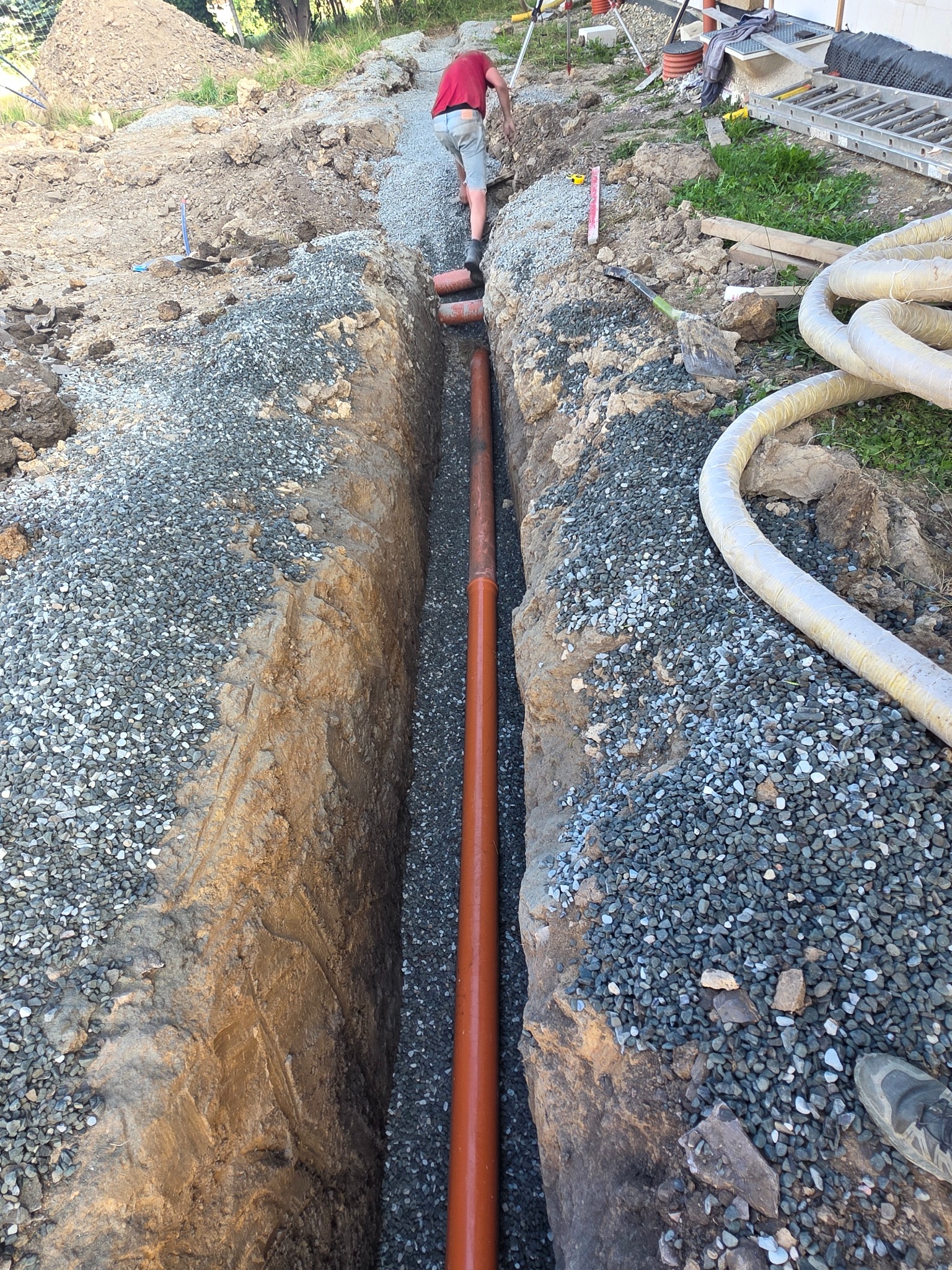 Wykop z rurą drenażową, obsypany żwirem. Widoczny fragment terenu z osobą pracującą w tle. Ujęcie z góry, ukazujące detale wykopu i materiałów.