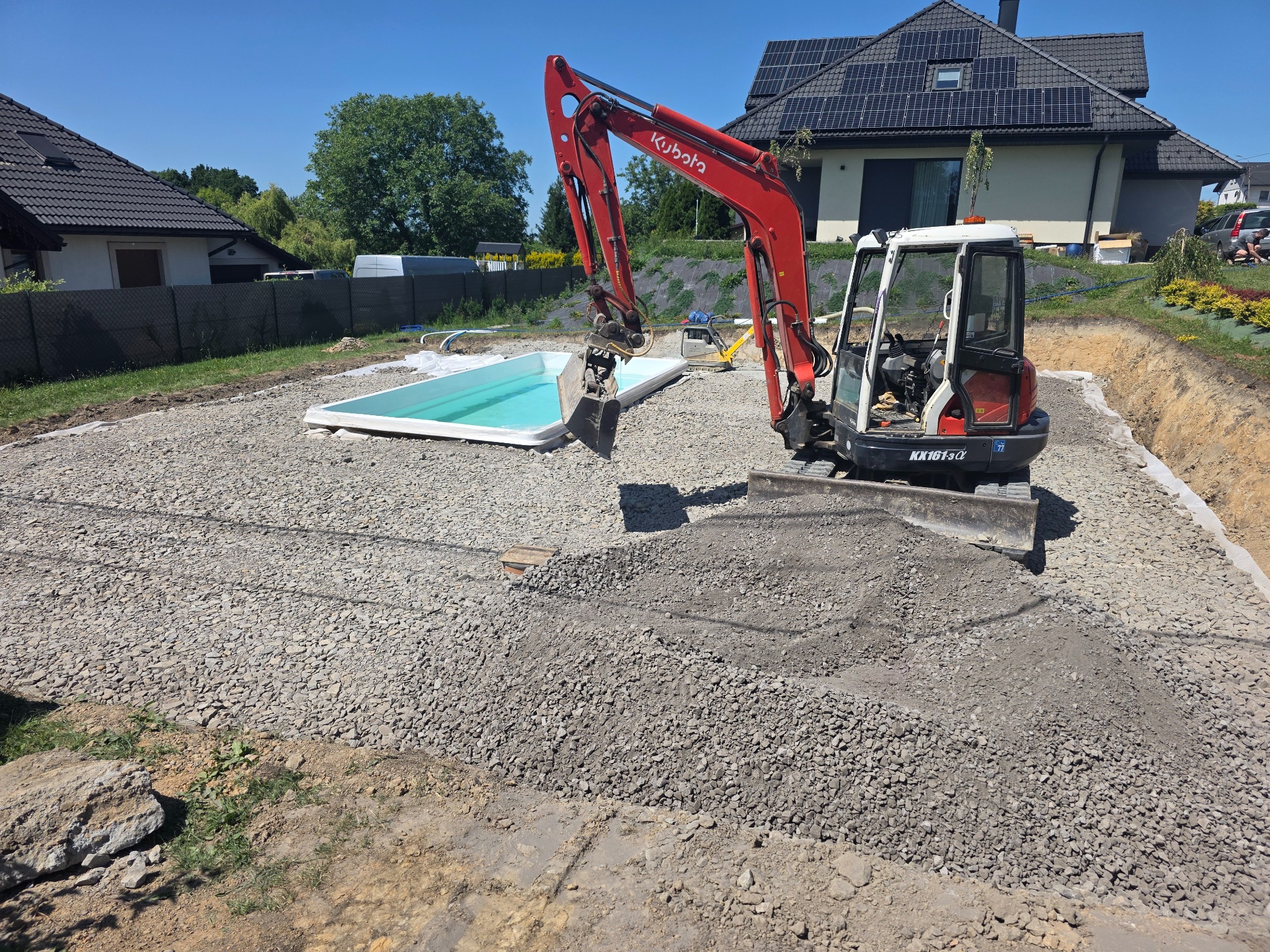 Czerwona mini koparka Kubota na żwirowym podłożu obok wkopanego basenu. W tle dom z panelami słonecznymi i zieleń.